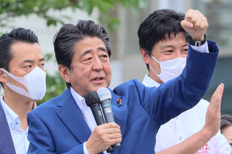 Abe speaking into a double microphone with his left arm raised in a fist; two men in N95 masks applaud behind him