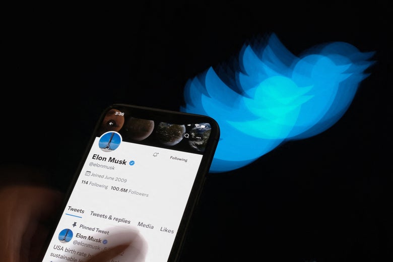 Elon Musk's Twitter profile is seen on a smartphone screen that is in turn held up against a background with a blurry Twitter bird.